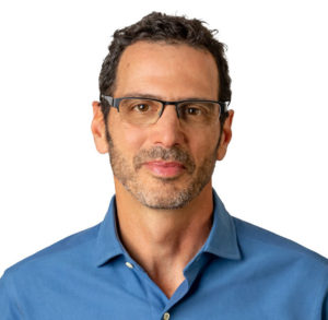 A man with short curly dark hair and glasses is wearing a blue collared shirt. He has a trimmed beard and is looking directly at the camera with a neutral expression. The background is plain white.
