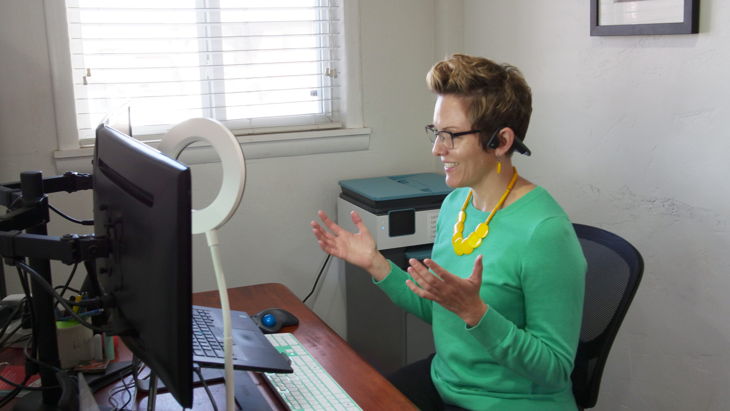 A person with short hair is sitting at a desk with a computer, web camera, and ring light. They are wearing glasses, a green sweater, and a yellow necklace, and are using a headset while gesturing with their hands, appearing to engage in a video call or virtual meeting.