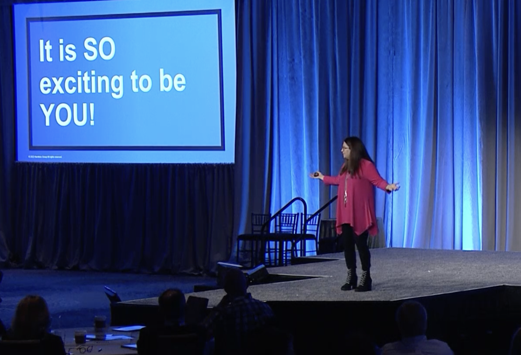 A woman in a bright pink top speaks onstage in front of a blue curtain. A large screen beside her displays the words, “It is SO exciting to be YOU!”.