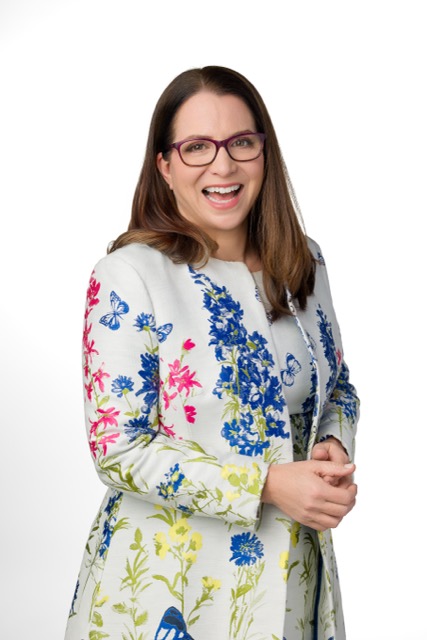 Williamson-441 Medium A woman with long brown hair and glasses smiles while standing against a white background. She wears a light-colored dress with colorful floral and butterfly patterns.