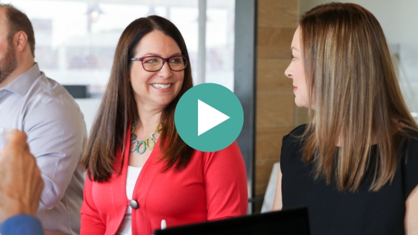 Two women sit together at a table in an office setting, smiling and talking. One woman wears glasses and a red cardigan; the other has straight blond hair and wears a black top. A play button icon overlays the image.