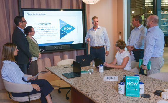 Six people are in a meeting room. One man stands by a screen giving a presentation, while five others sit or stand around a table with laptops and papers, attentively listening and watching the screen.