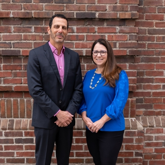 A man in a dark suit and pink shirt stands next to a woman in glasses and a bright blue blouse, both smiling, in front of a red brick wall.