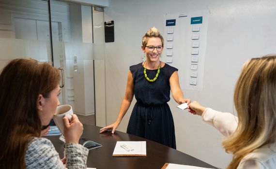 A woman standing and smiling hands a business card to another woman seated at a conference table, while a third woman with a coffee cup listens. A whiteboard with notes is visible in the background.