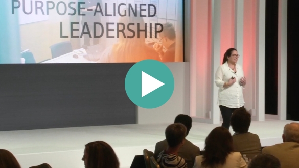 kg-video-sizzle-reel A woman stands on stage giving a presentation titled "Purpose-Aligned Leadership" to an audience; a large screen displays the presentation title behind her.