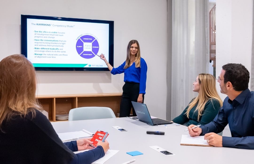 A woman stands by a screen giving a presentation to three seated colleagues in a meeting room. She points at a slide with a circular diagram while others listen and take notes.