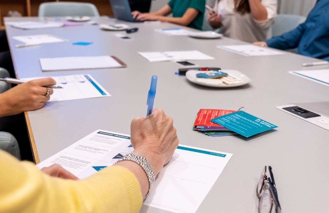 People sit around a conference table with papers, pens, and informational cards. One person writes while others discuss. Coffee cups, glasses, and folders are also visible on the table, suggesting a meeting or workshop setting.