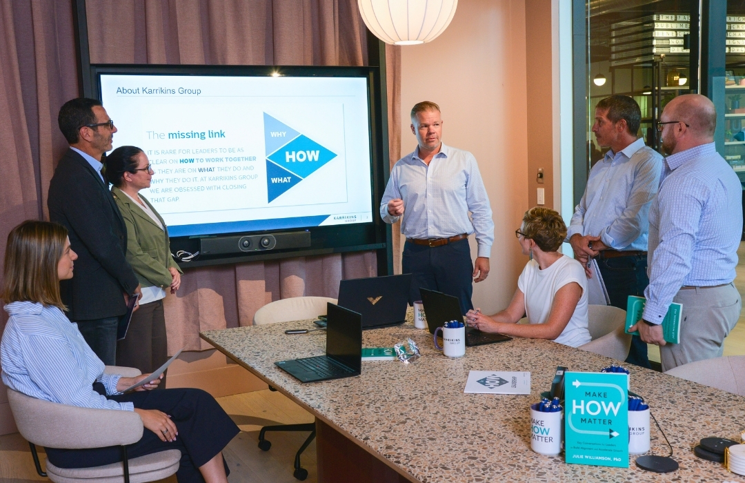 kg-enterprise-team-alignment Six professionals have a meeting in a modern office. One man stands by a screen giving a presentation about “The missing link.” Others sit or stand, listening, with laptops, notebooks, and branded mugs on the table.