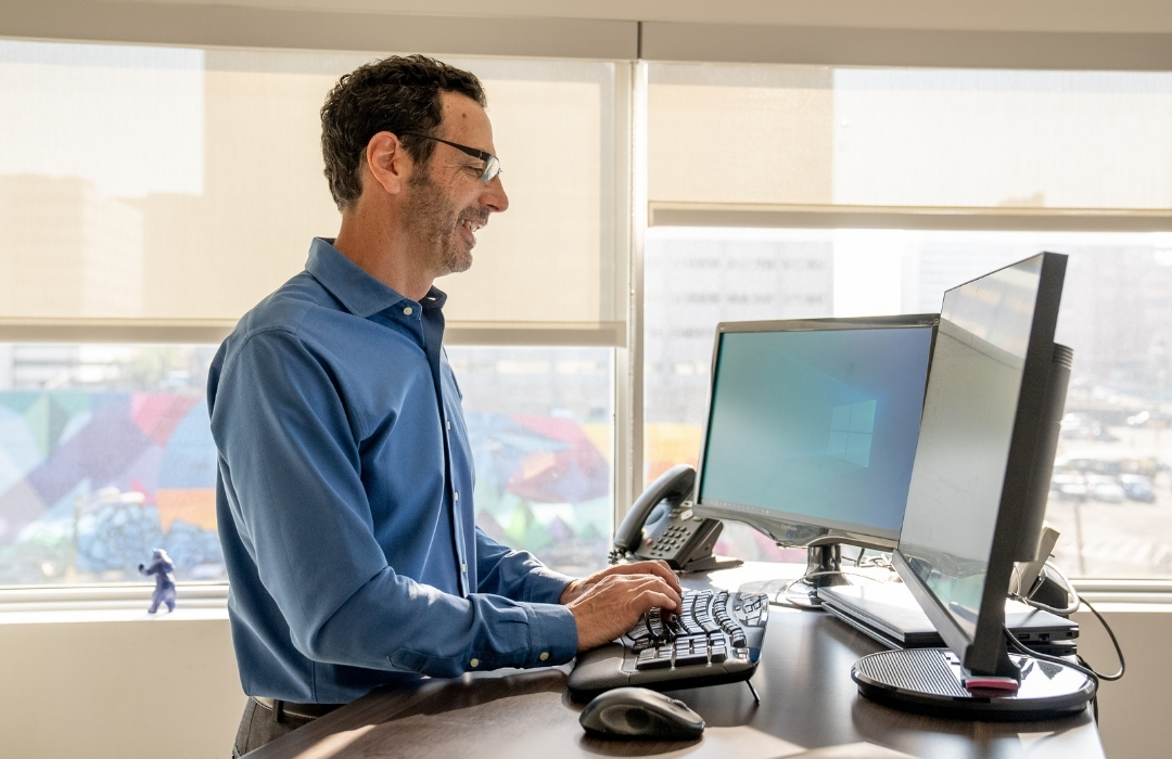 kg-executive-coaching Man in blue shirt standing at a desk, typing on a keyboard and looking at dual monitors in a bright office with large windows and cityscape outside.