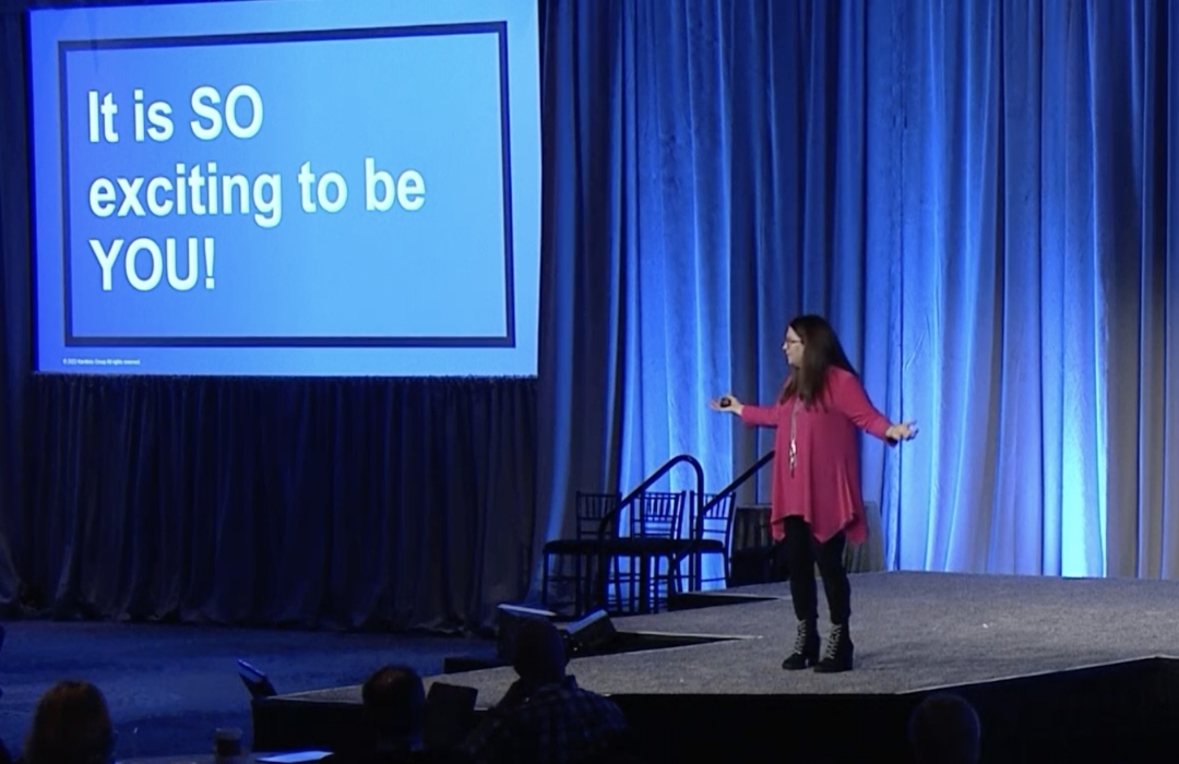 kg-public-speaking A woman in a pink top stands on a stage next to a screen that reads, “It is SO exciting to be YOU!” Blue curtains hang in the background.