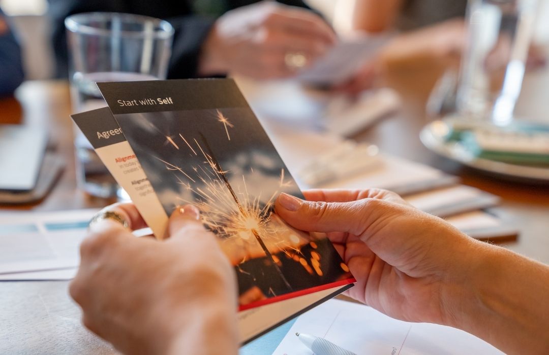 A person holds a brochure titled “Start with Self” showing a sparkler, with other people and documents blurred in the background on a table.